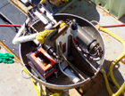 Top view of release module.  Components are housed in a 60? diameter stainless steel elliptical dish.  Modem electronics, hi-powered battery pack and retracted altimeter boom are visible.  