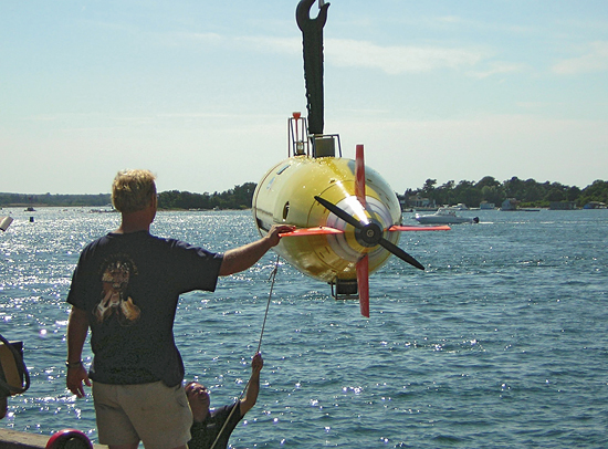 At the WHOI dock, Aaron Kayes of the Naval Oceanographic Office (NAVOCEANO) holds the rope line and Frank Raspante of Hydroid, Inc., holds a tail fin as the REMUS-6000 vehicle is lowered into the water for trials and testing. The autonomous underwater vehicle?designed to reach depths of 6,000 meters?was developed and built by the WHOI Oceanographic Systems Lab for NAVOCEANO. 