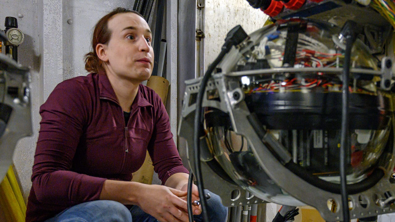 WHOI mechanical engineer Casey Machado sits in the ship’s staging bay before Orpheus’ first plunge of the expedition. The vehicle’s electronics are housed in a glass sphere, shown in the foreground, which can tolerate the extreme pressure of the deep, and is one of many cost-saving measures that went into the vehicle’s design. (Photo by Evan Lubofsky, Woods Hole Oceanographic Institution)