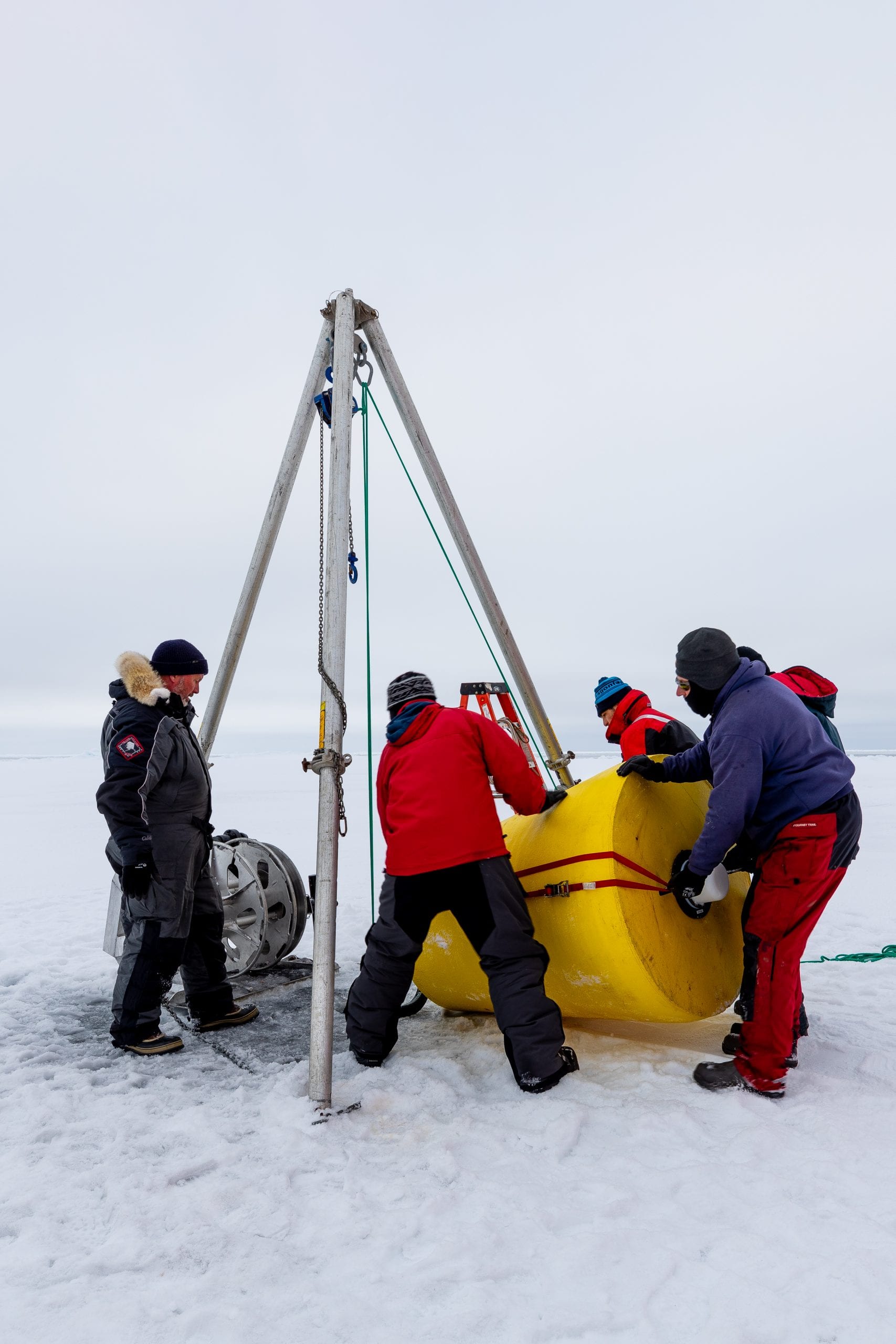 Four people position a yellow mooring under a metal gantry positioned over a hole in the sea ice.