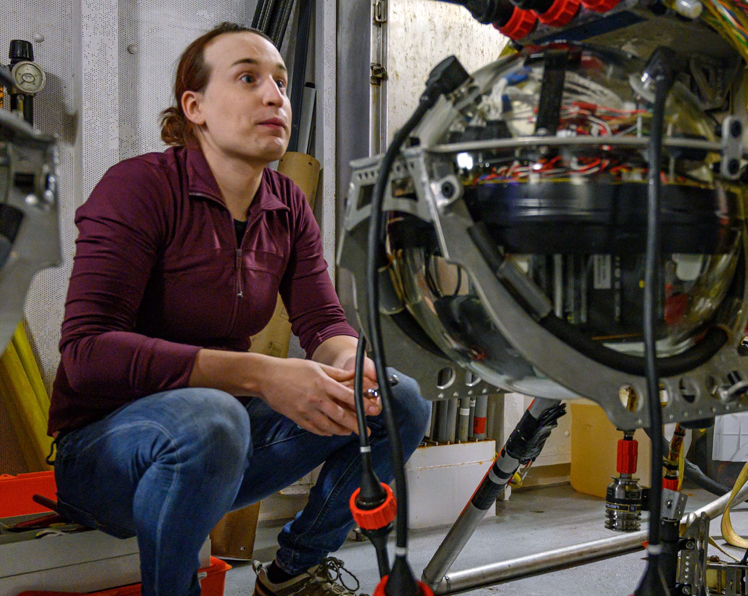 WHOI mechanical engineer Casey Machado sits in the ship’s staging bay before Orpheus’ first plunge of the expedition. The vehicle’s electronics are housed in a glass sphere, shown in the foreground, which can tolerate the extreme pressure of the deep, and is one of many cost-saving measures that went into the vehicle’s design. (Photo by Evan Lubofsky, Woods Hole Oceanographic Institution)