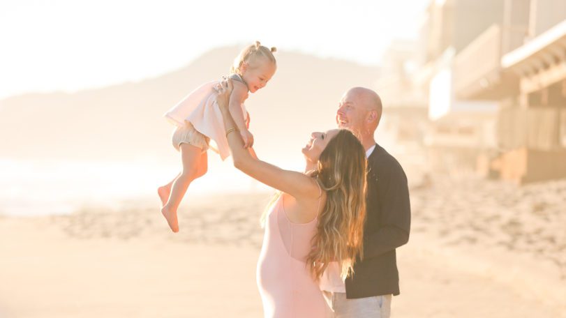 A pregnant woman in a pink dress lifts a female toddler in a pink dress while a man in a black coast and white pants smiles at them. They are on a sunny beach with buildings, mountains and the ocean in the background.