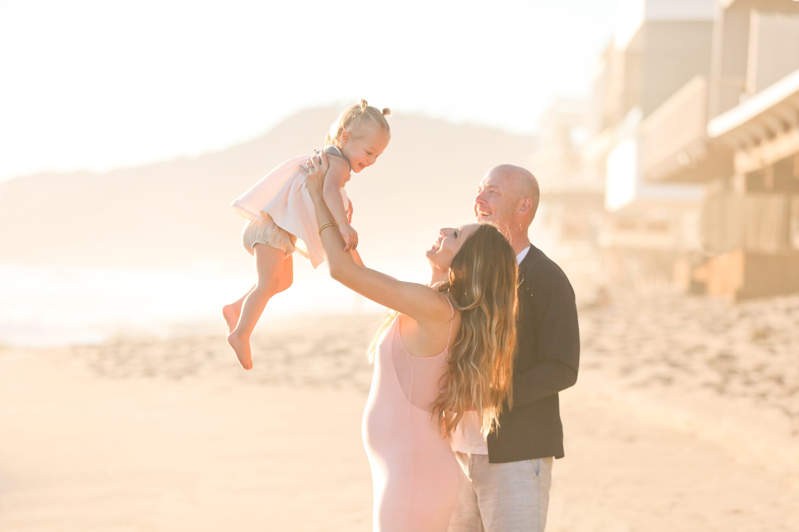 A pregnant woman in a pink dress lifts a female toddler in a pink dress while a man in a black coast and white pants smiles at them. They are on a sunny beach with buildings, mountains and the ocean in the background.