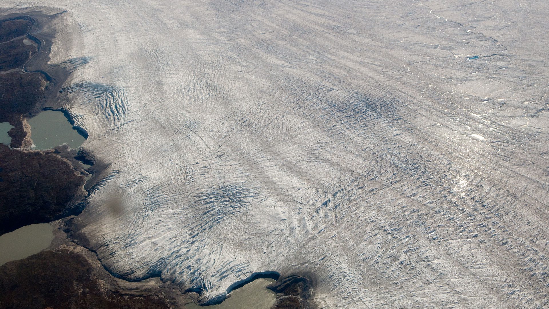 From the air, Greenland's ice sheet looked like white molasses oozing down the mountainside and into the sea. Researchers are investigating Greenland's glacial lakes, which form atop the ice sheet each spring and summer as returning sunlight melts ice and snow. They have found that as lake grow larger, large cracks can open up at their bases, allowing the lake water to drain in a dramatic surge all the way to the bedrock at the bottom of glaciers. The water lubricates the base of the glacier, like grease on a railroad track, allowing glaciers to flow faster toward the coast and discharge more ice to the ocean