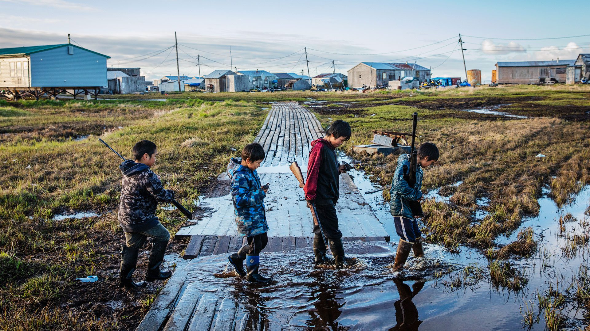 Four boys cross a flooded walkway in the Yip’Ik Village of Newtok, Alaska following a recent bird hunt. They are among the residents now relocating as melting permafrost causes their land to sink between two major rivers and the sea.
