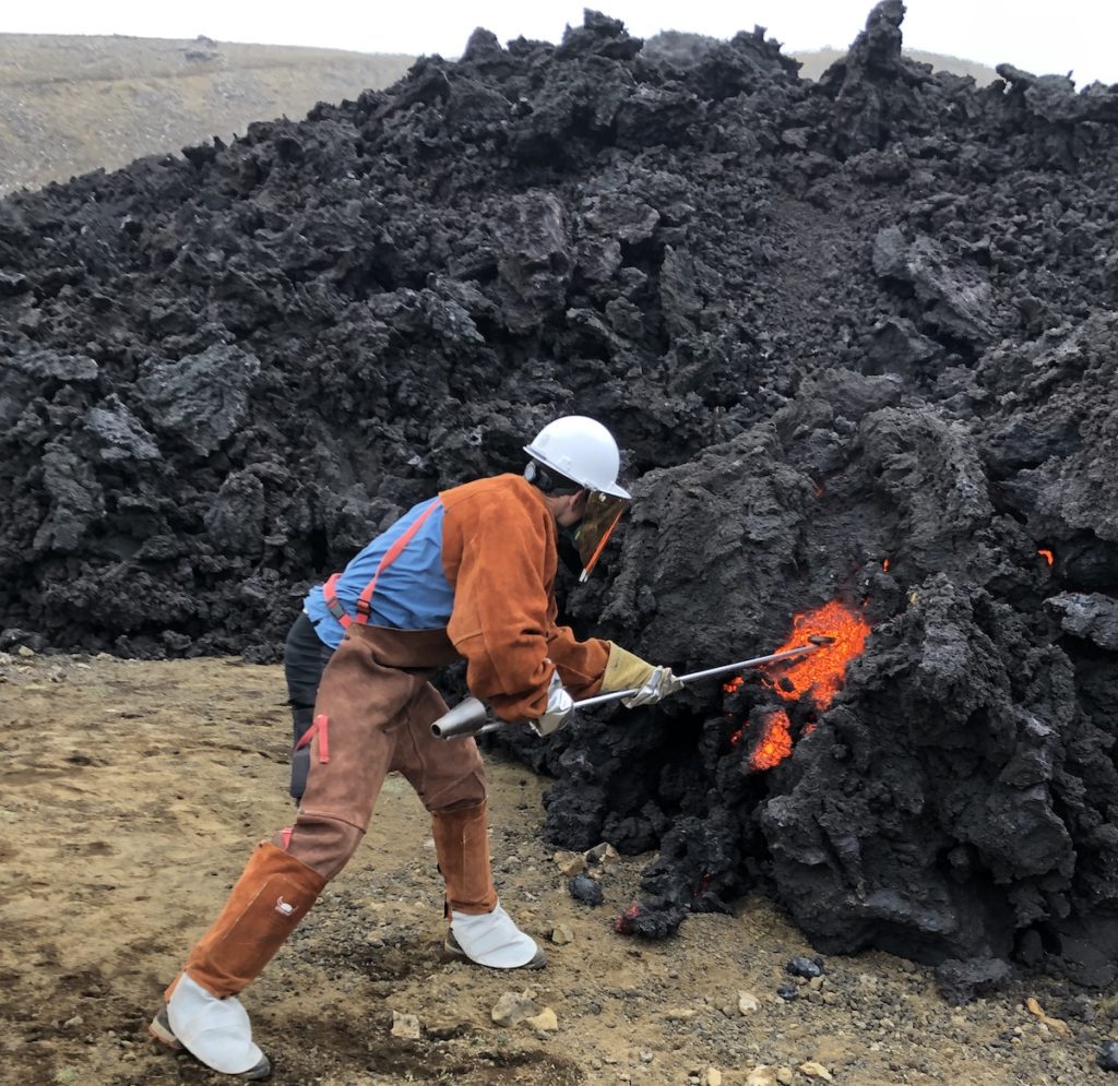 A man dressed in orange samples orange lava from black volcanic rock.