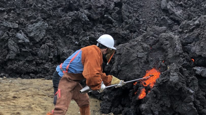 A man dressed in orange samples orange lava from black volcanic rock.