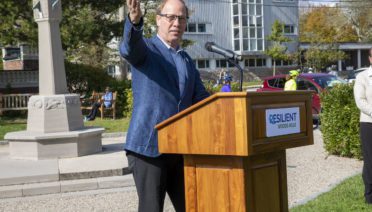 A man with glasses and a blue suit stands at a podium with the words Resilient Woods Hole on it. He's gesturing to his right and the Woods Hole sundial and MBL buildings are in the background.