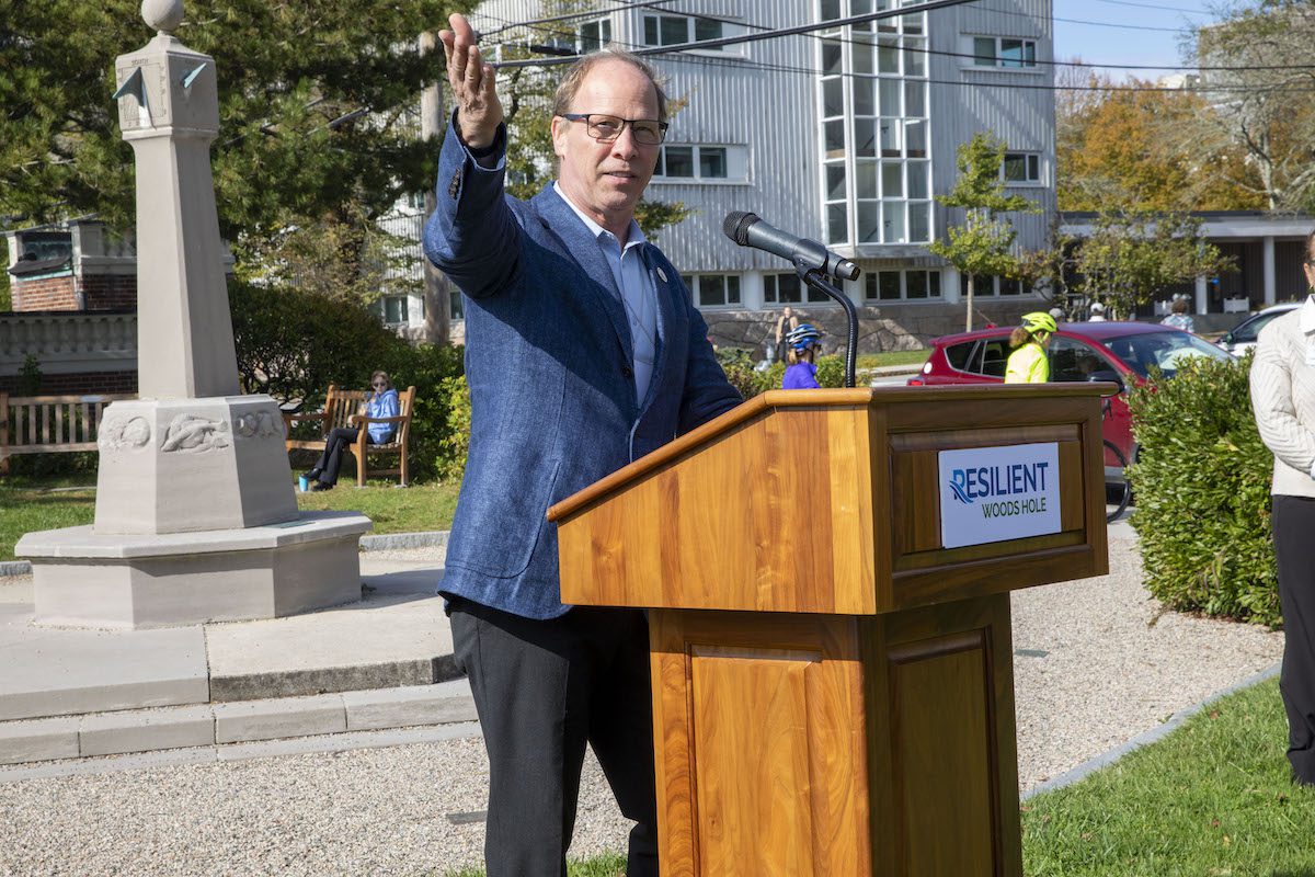 A man with glasses and a blue suit stands at a podium with the words Resilient Woods Hole on it. He's gesturing to his right and the Woods Hole sundial and MBL buildings are in the background.