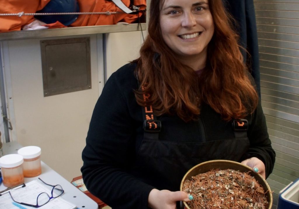 A smiling white young woman with brown hair holds a round metal container containing assorted orange sea life. Starfish are visible.